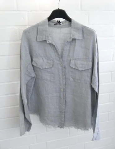 Hellgraue Damen Leinenbluse mit Brusttaschen und ausgefranstem Saum vor weißer Wand.
